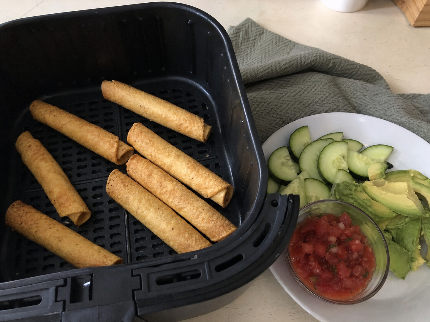 Crisp taquitos spaced in an air-fryer basket beside avocado, cucumber and salsa