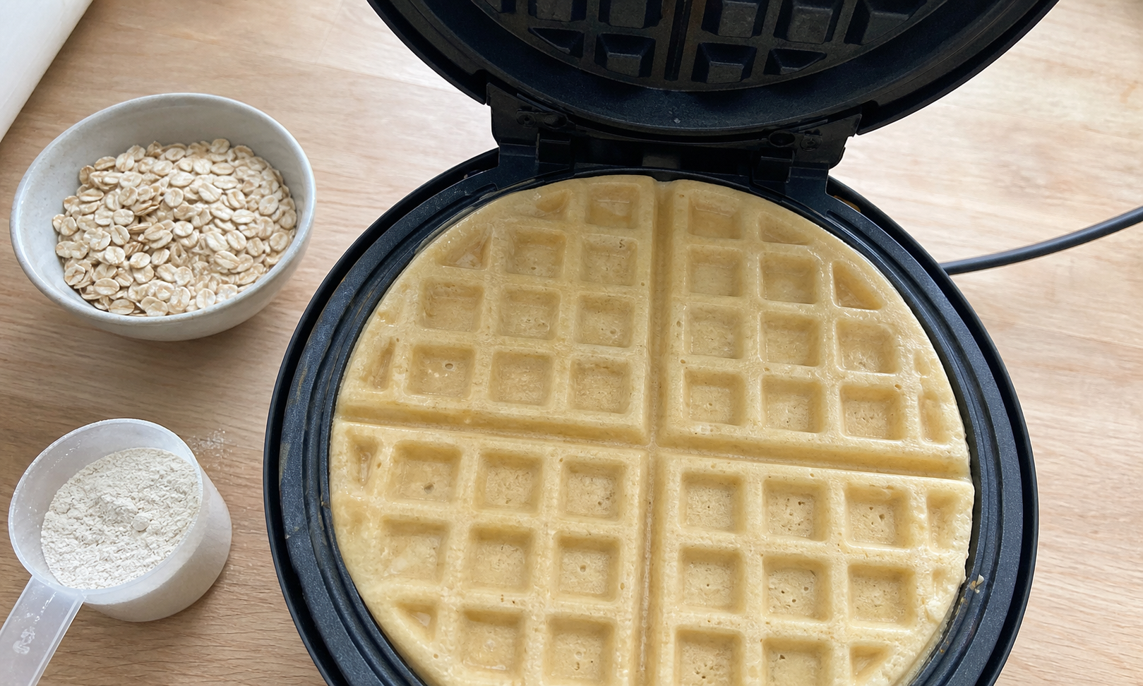Protein waffle cooking in an open waffle maker beside oats and protein powder