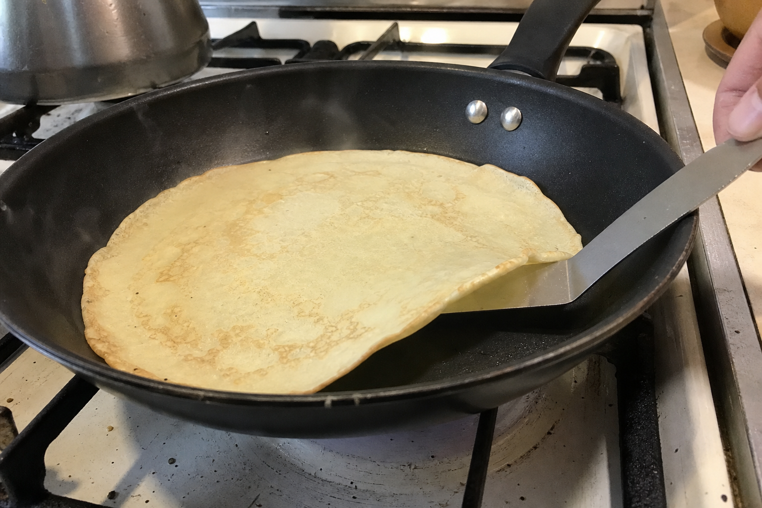 A thin crepe warming in a skillet while a spatula lifts the edge